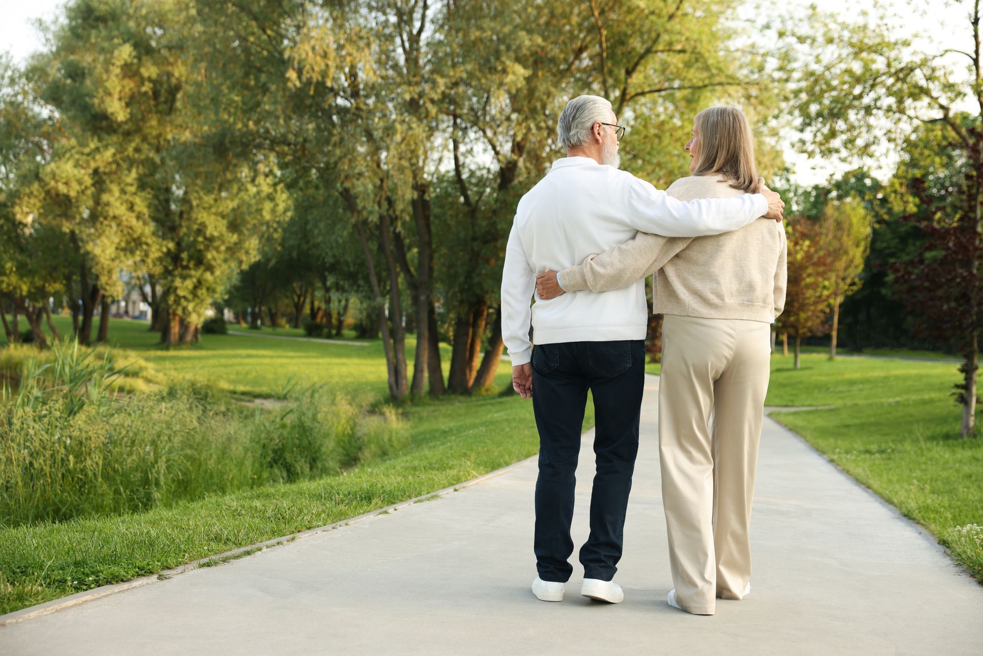 Two people walking arm in arm on a sunlit park path, surrounded by green trees and grass.