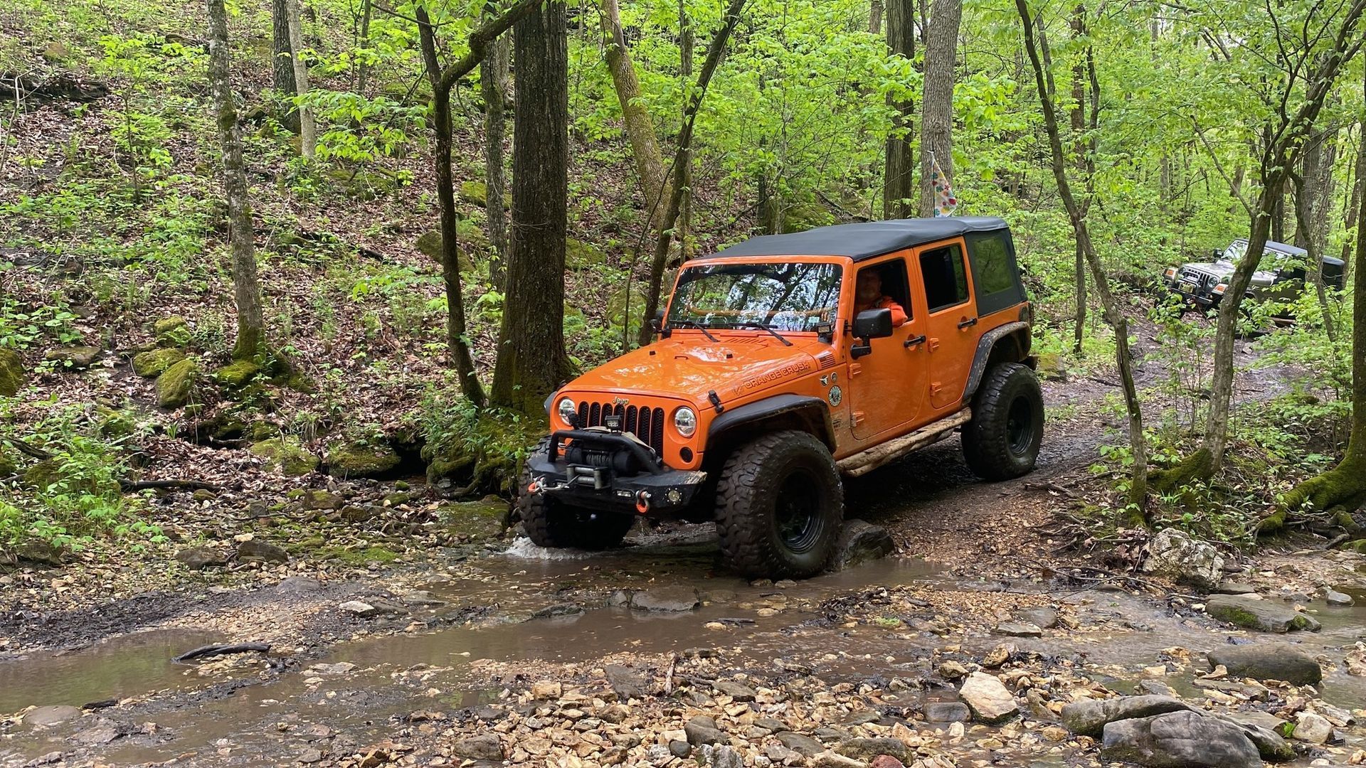 Orange Jeep Wrangler driving through a muddy forest path.