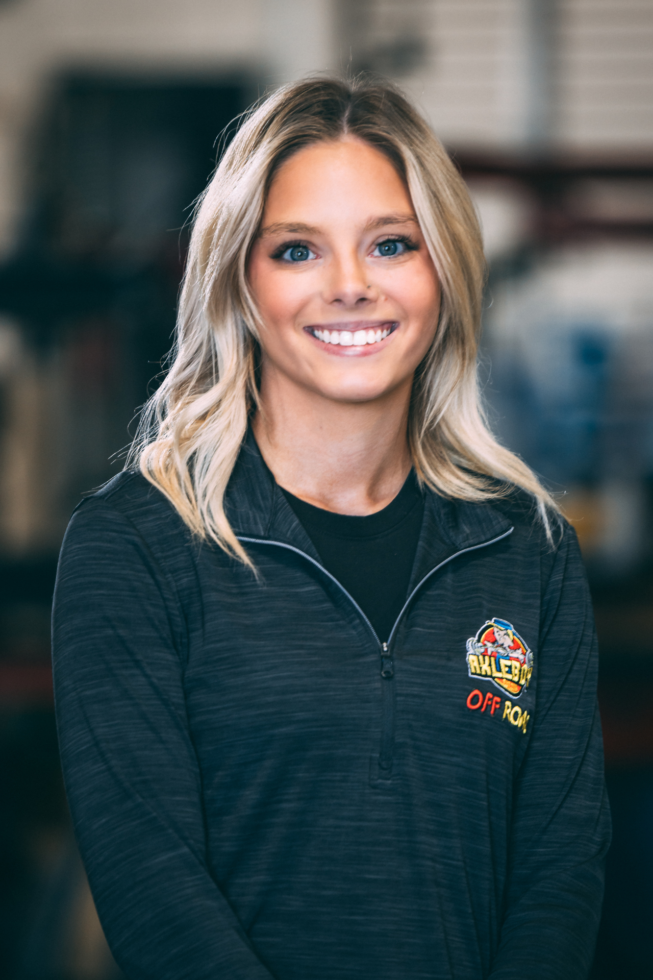 Blonde woman smiling, wearing a black long-sleeved shirt with logos. Indoors, blurred background.