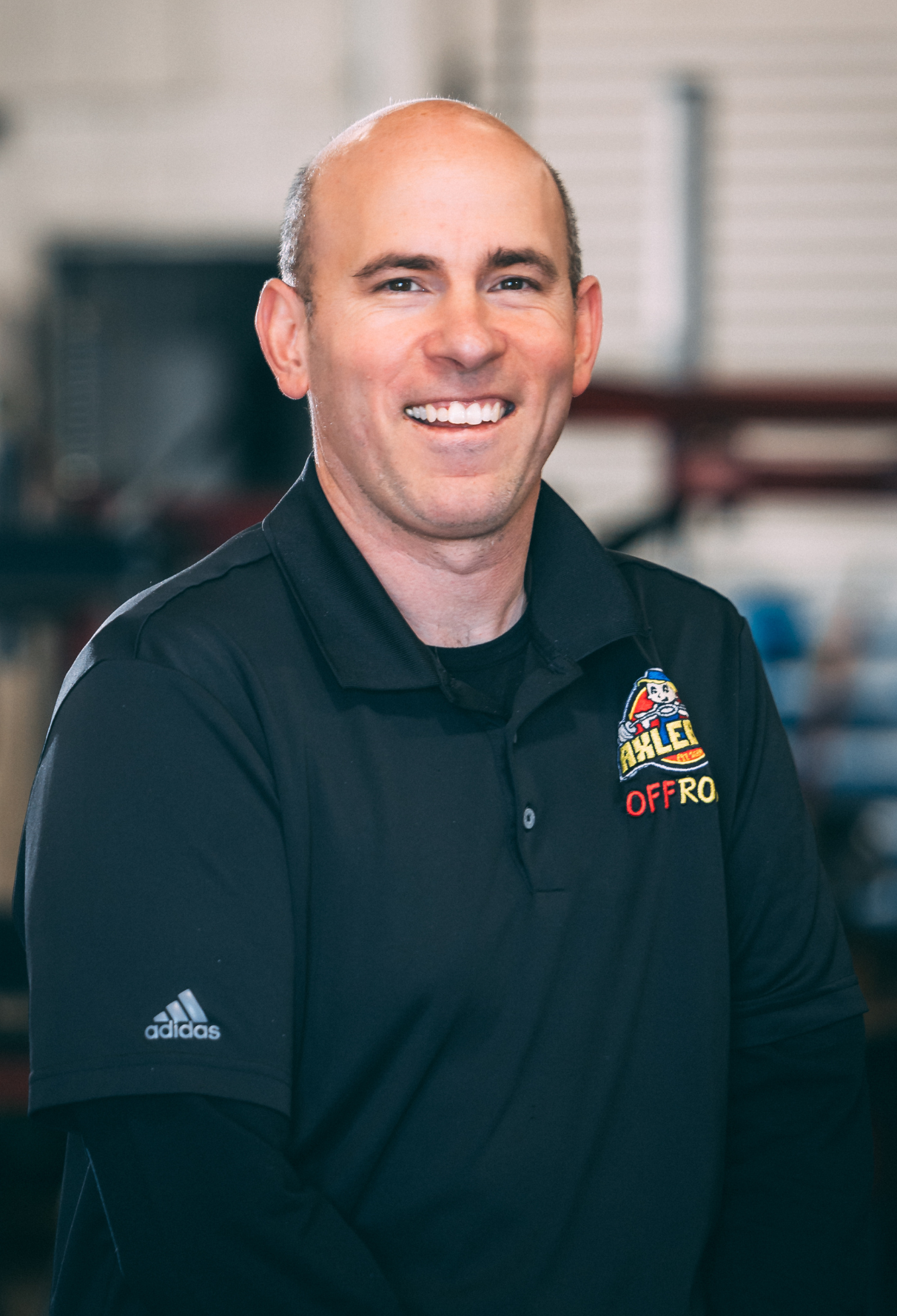 Bald man wearing a black polo shirt with a logo, smiling at the camera in a shop setting.