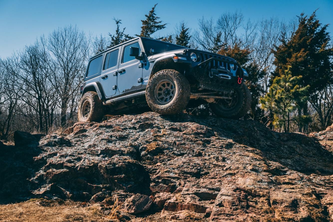 Blue Jeep Wrangler off-roading on a rocky terrain, with trees in the background on a sunny day.