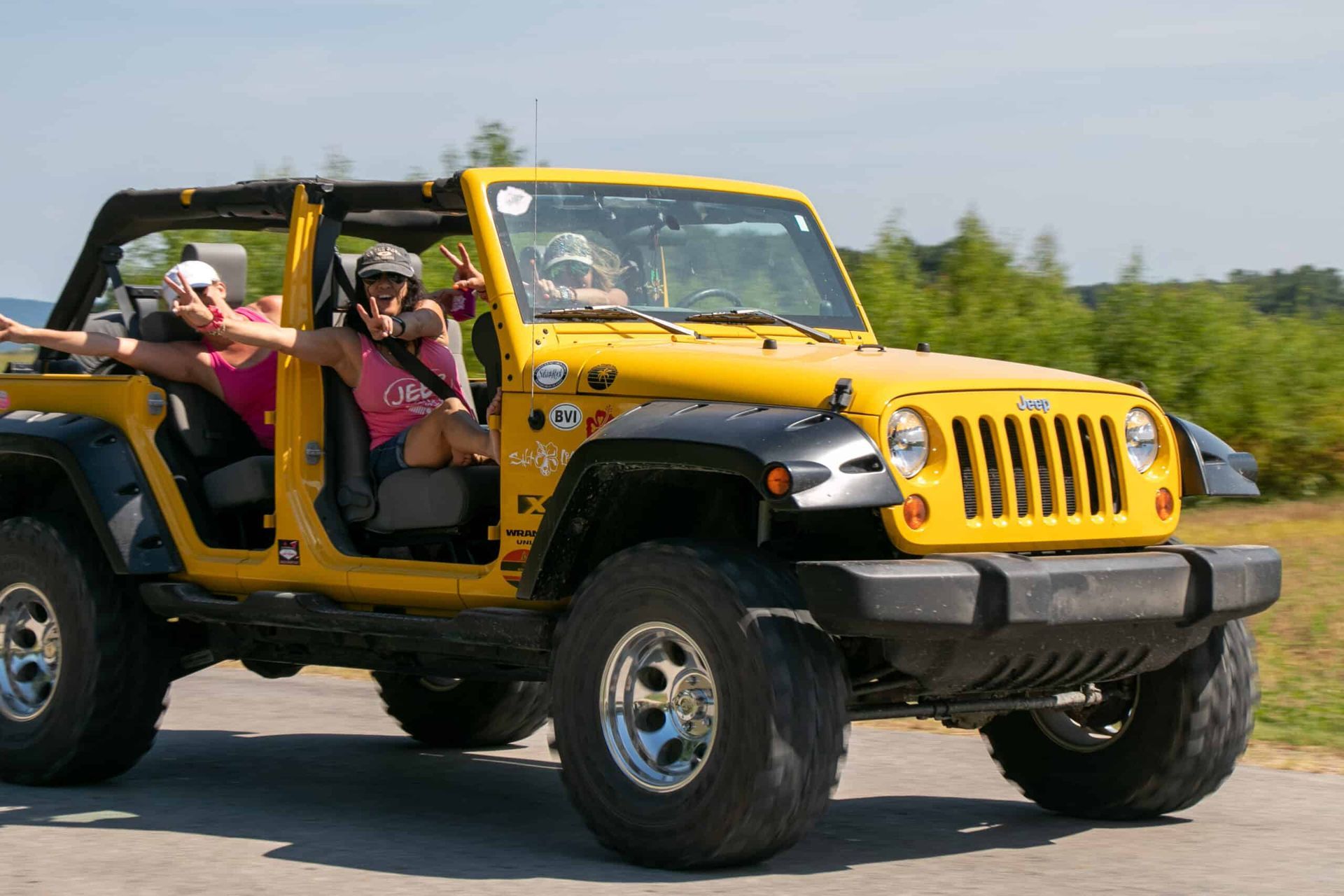 Yellow Jeep with three people, waving and smiling, driving on a road in a sunny, outdoor setting.