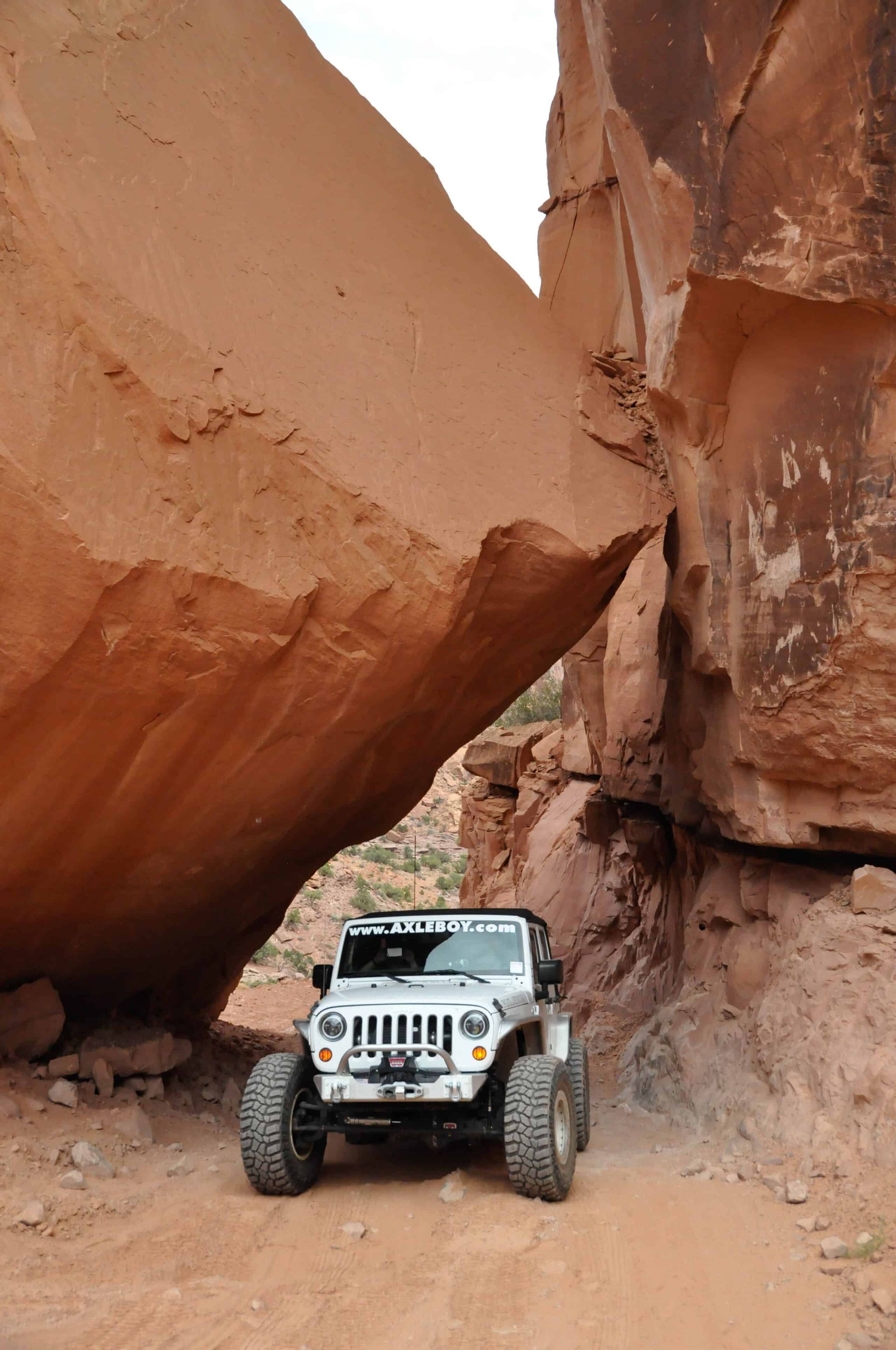 White Jeep on a dirt road under a large red rock formation in a desert environment.