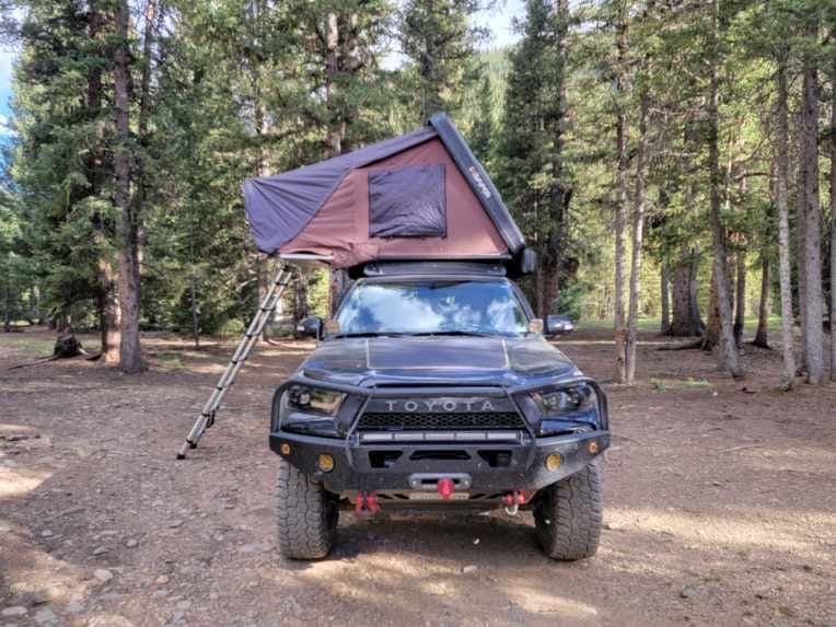 Black Toyota truck with rooftop tent, parked in a forest campsite.