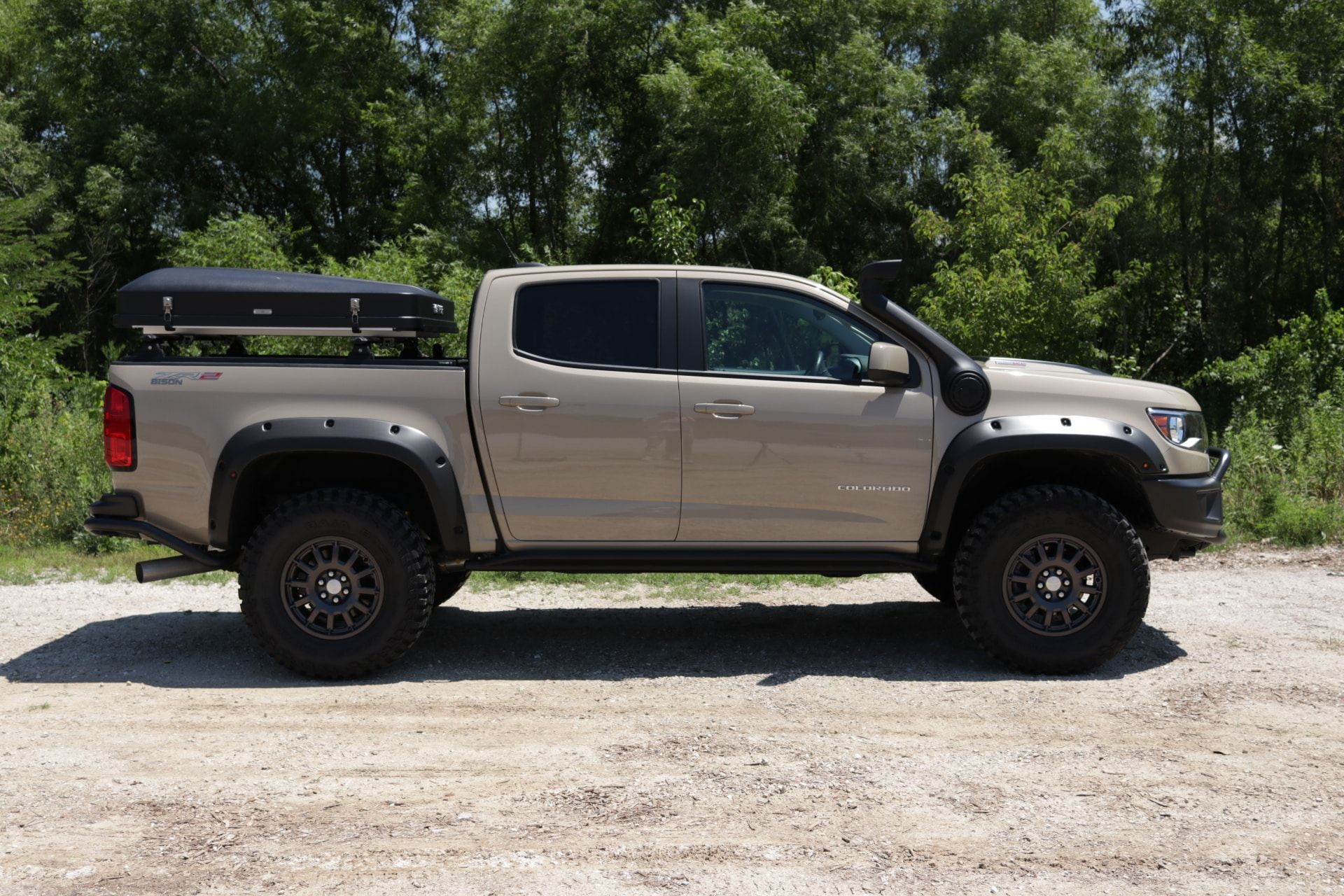 Tan lifted pickup truck with a snorkel, black fender flares, and rooftop tent on a gravel road.