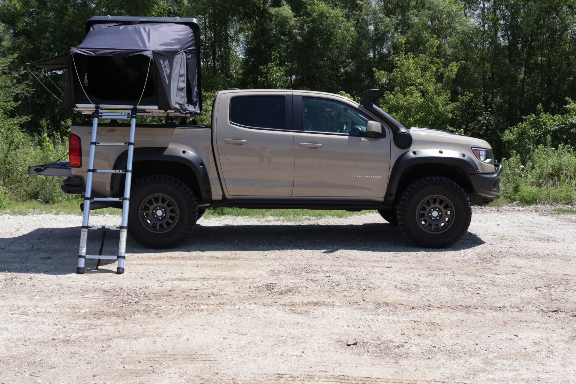 Tan pickup truck with rooftop tent, ladder extended on gravel road.