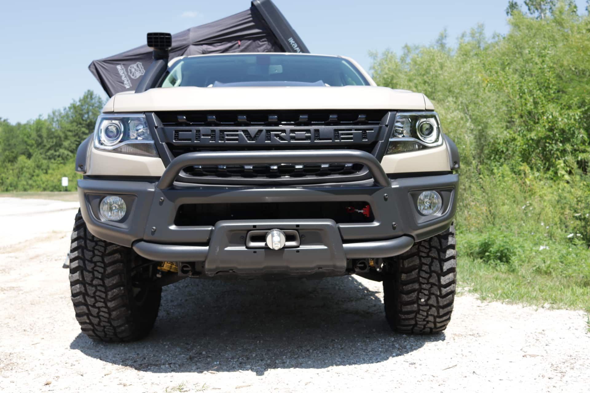 Tan Chevrolet pickup truck with off-road tires, front bumper, and a rooftop tent parked outdoors.