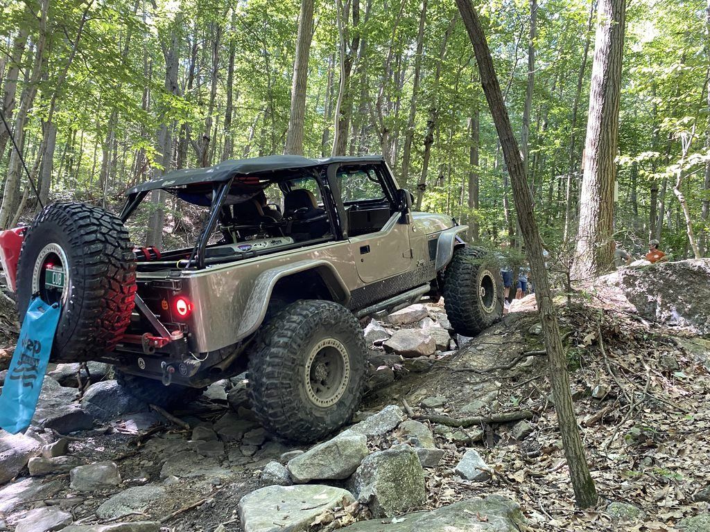 Tan Jeep off-roading on a rocky trail in a forest.