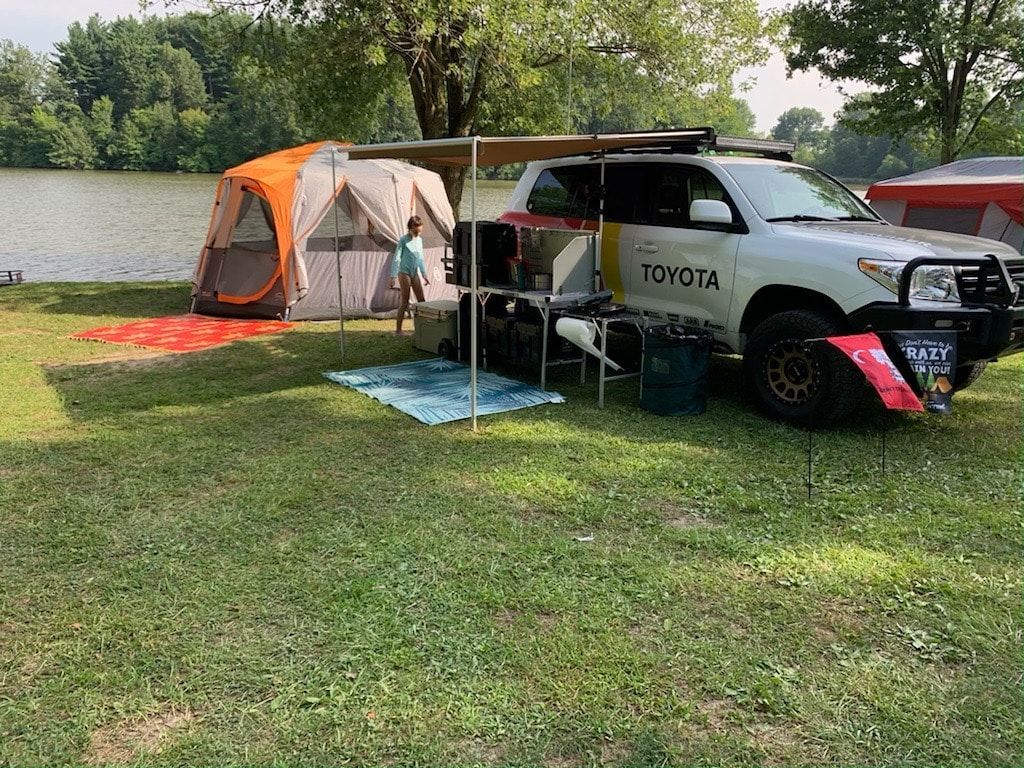 A white Toyota truck set up for camping next to a lake, with a tent and awning.