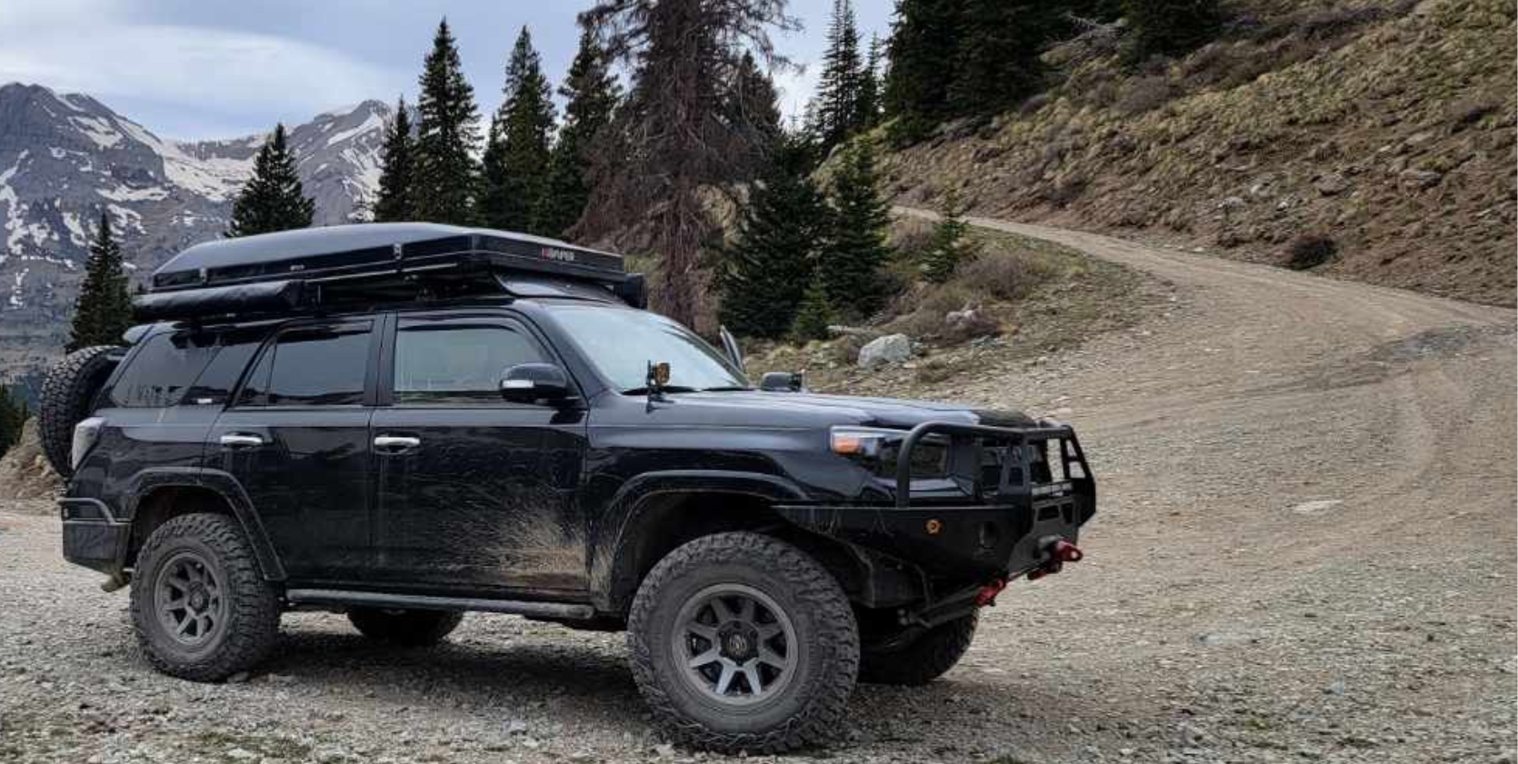 Black SUV on a dirt road in a mountain setting. It has a roof rack and is covered in mud.