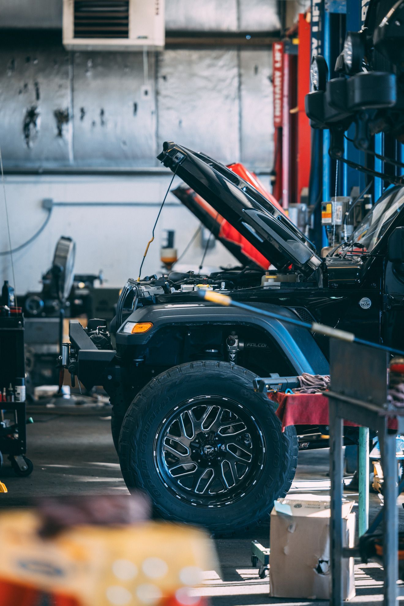 Jeep Wrangler with hood open in a garage. Black with large wheels, blue accents, red lift.