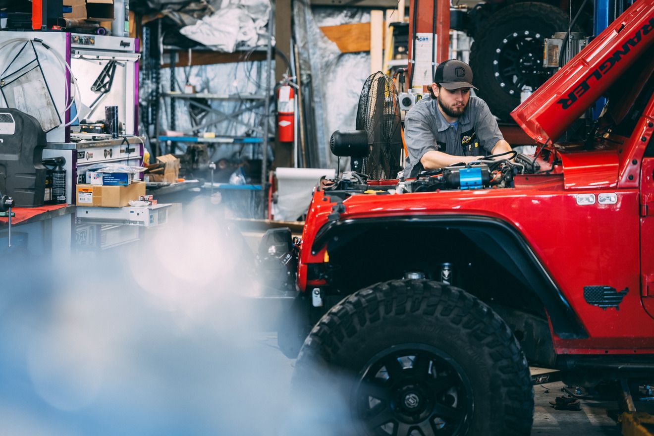 Mechanic working on a red Jeep in a garage, hood open. Blue smoke billowing from the engine.