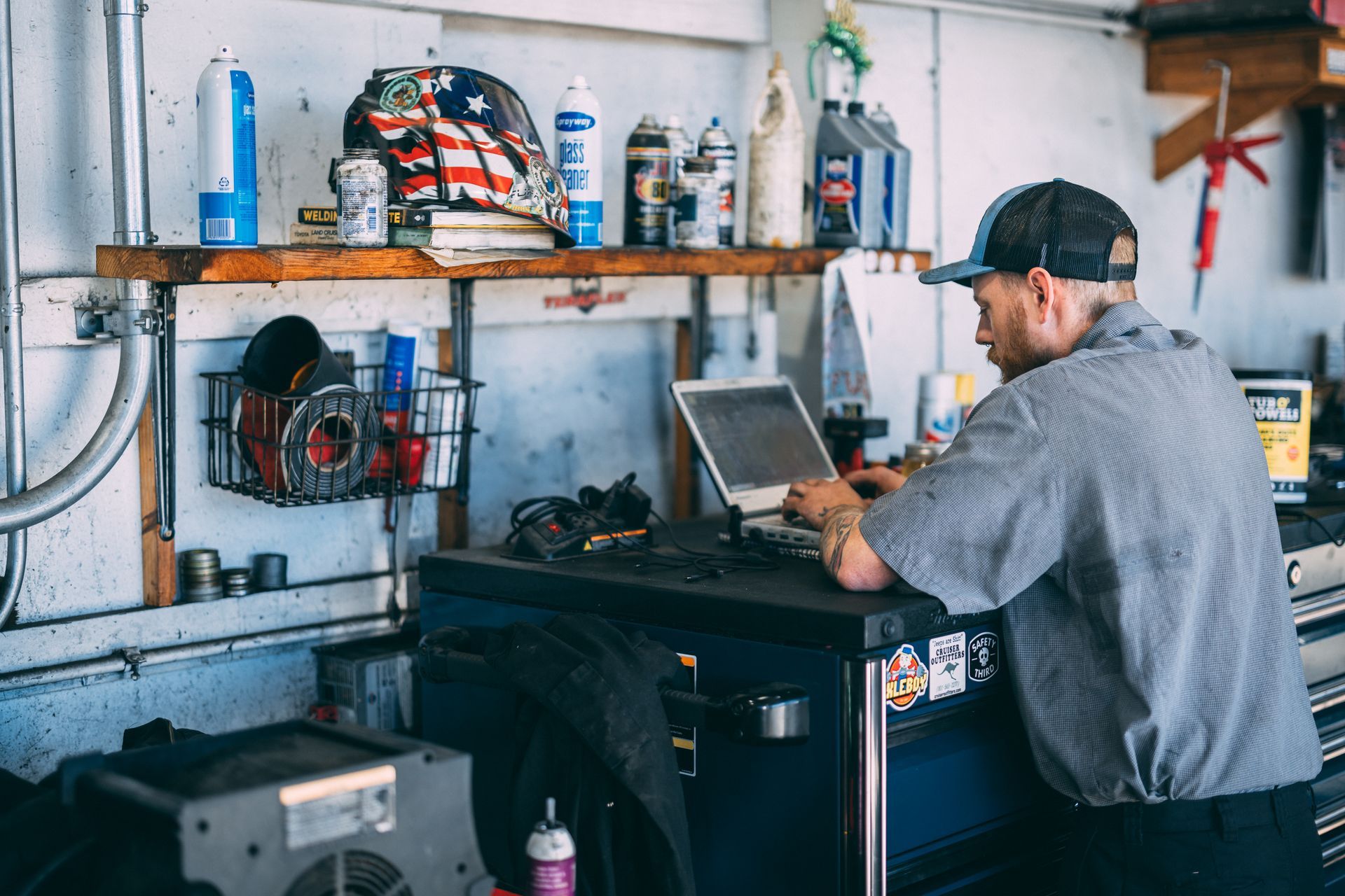 Mechanic in garage, working on laptop at a tool chest. A shelf holds various items.