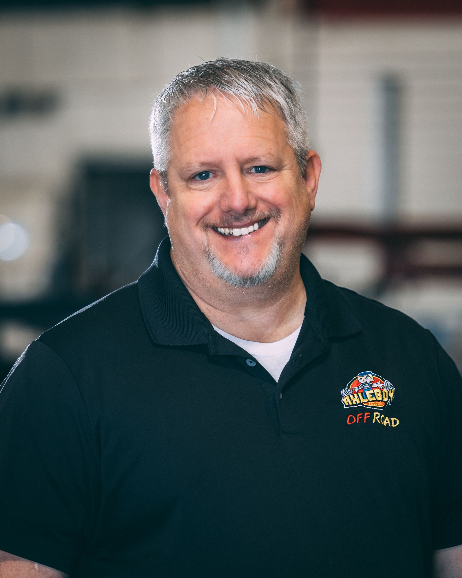 Smiling middle-aged man with graying hair wearing a black polo shirt, possibly in a workshop.