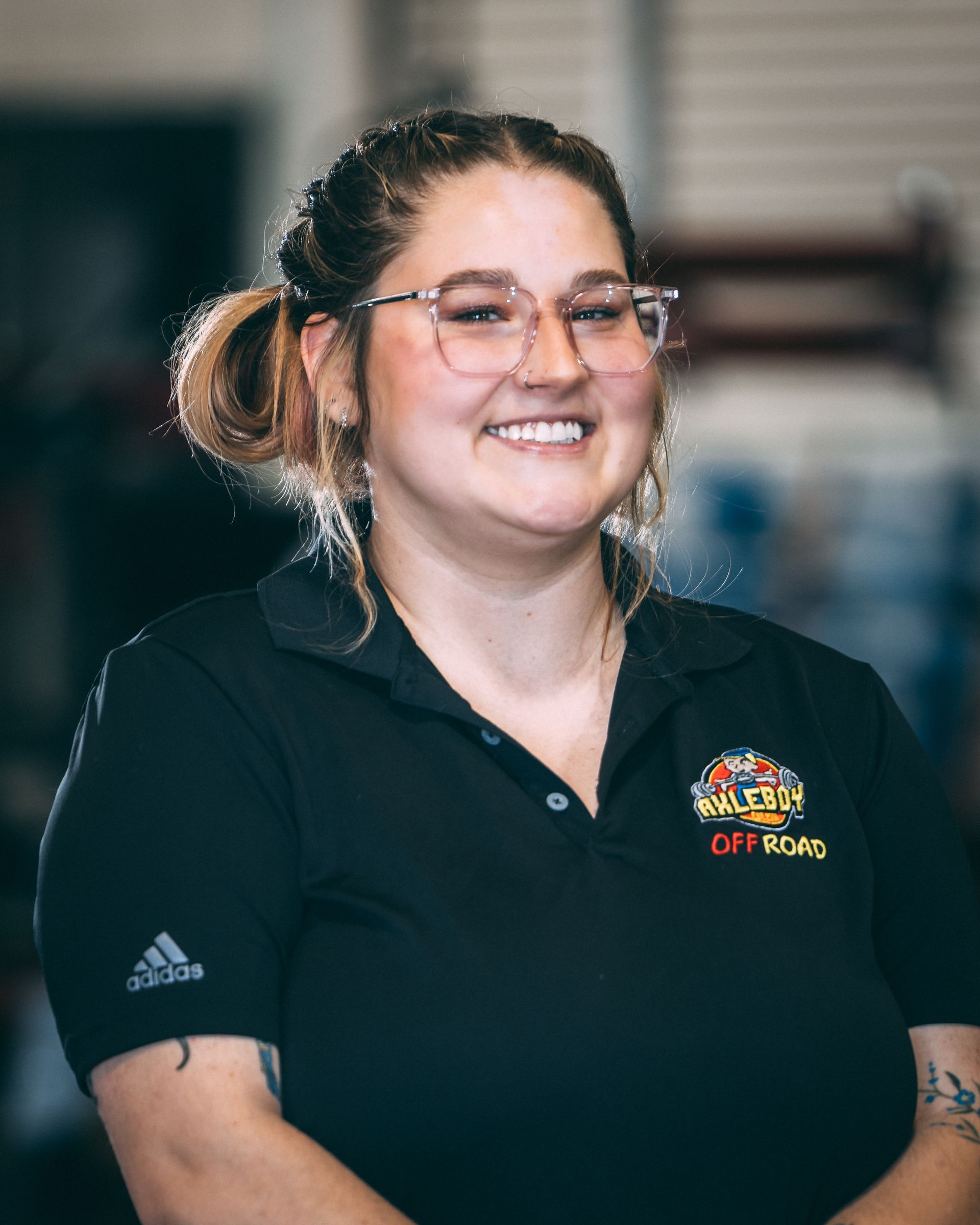 Woman with glasses, smiling, in black polo shirt with logo. Inside a workshop, smiling.
