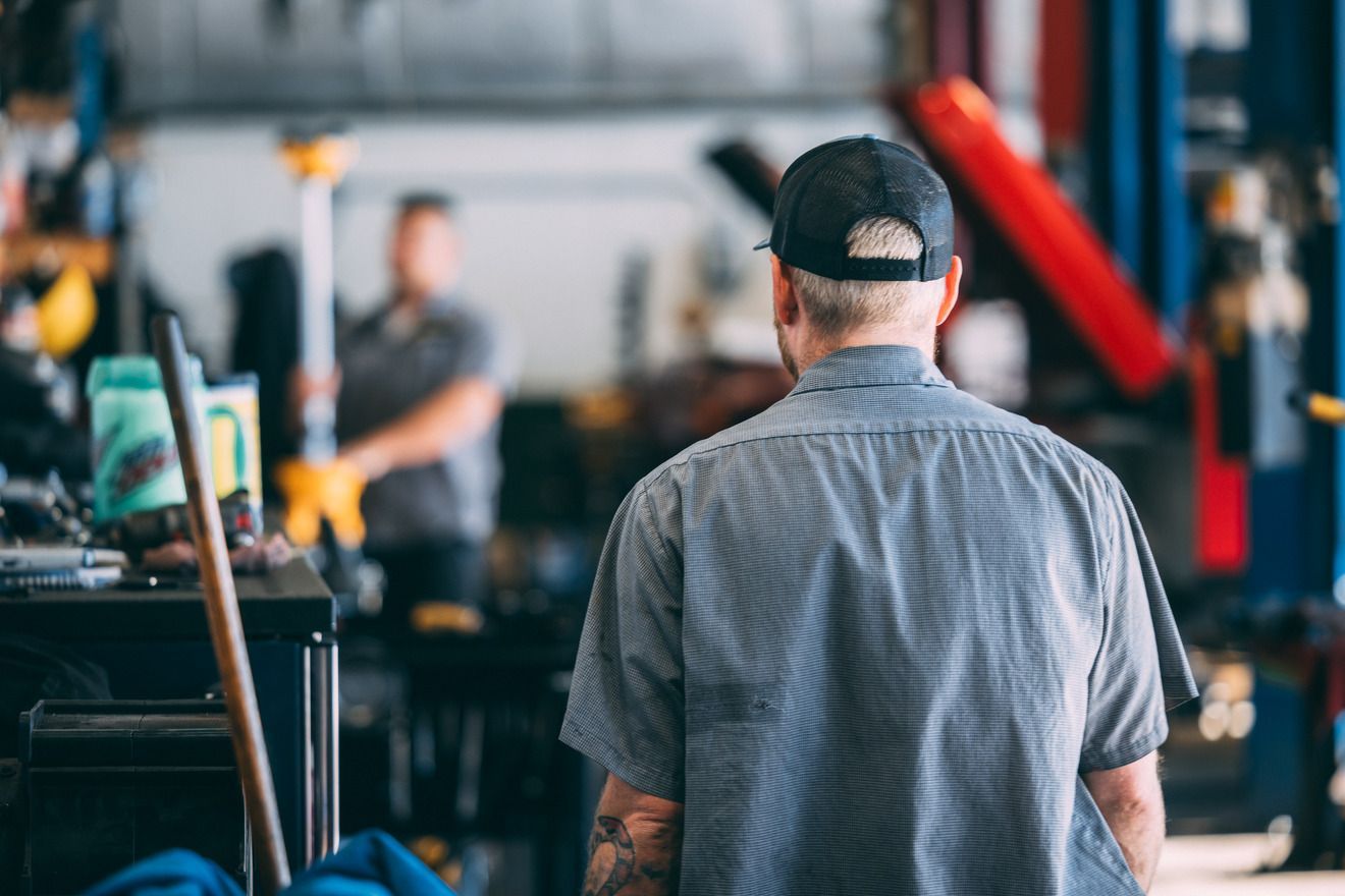 Mechanic in a gray shirt and cap in a garage, looking at another mechanic working in the background.