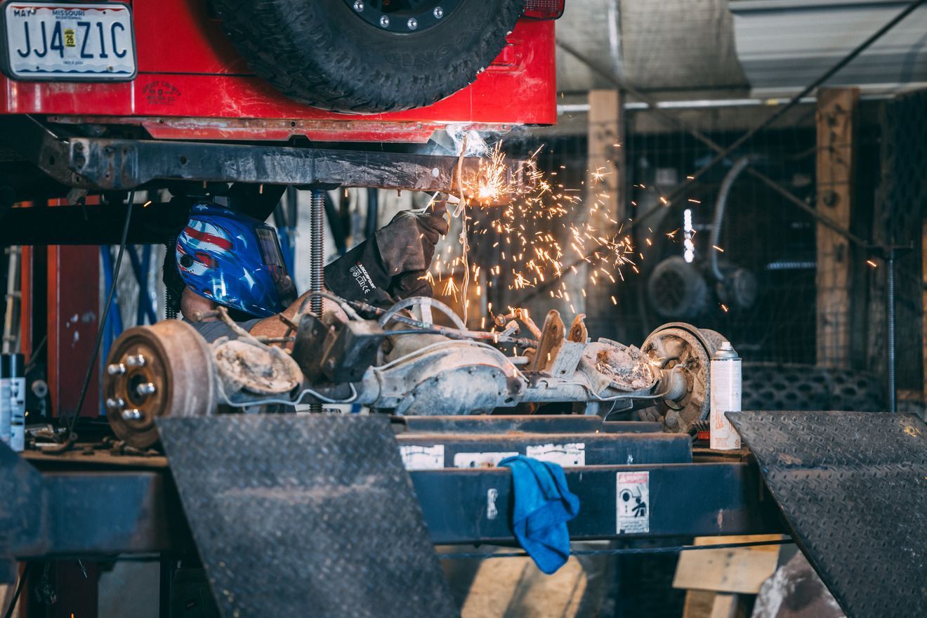 A mechanic welds under a red Jeep in a garage, sparks flying.