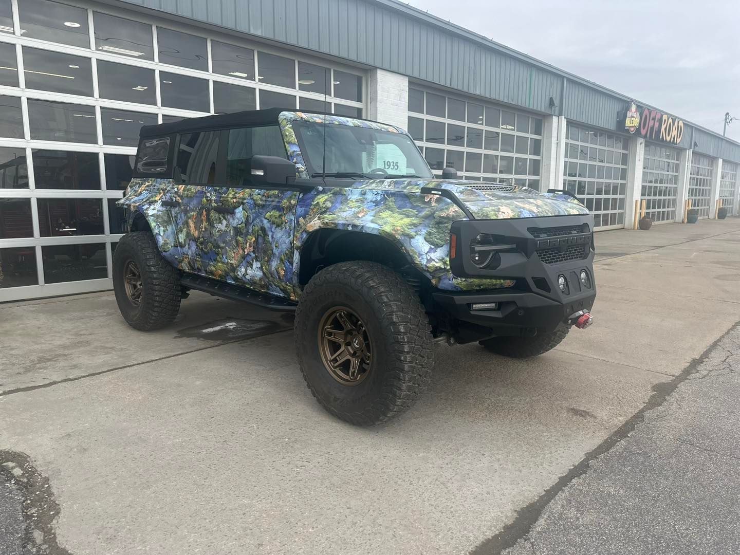 Camouflage-wrapped Ford Bronco parked outside a commercial building with garage doors.