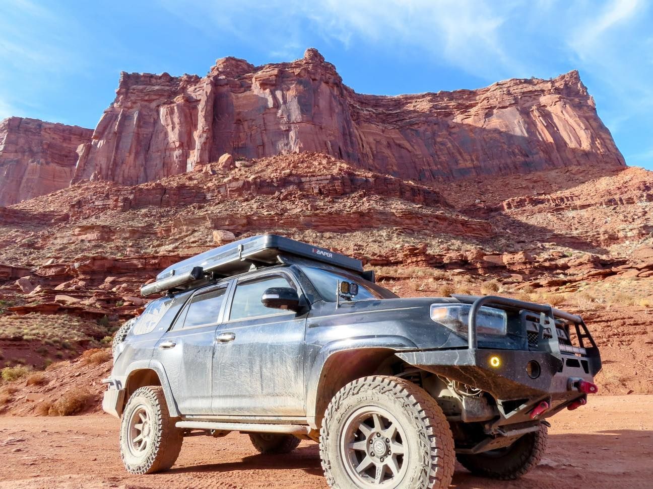 Black off-road SUV parked on dirt road, red rock cliffs in background, sunny day.