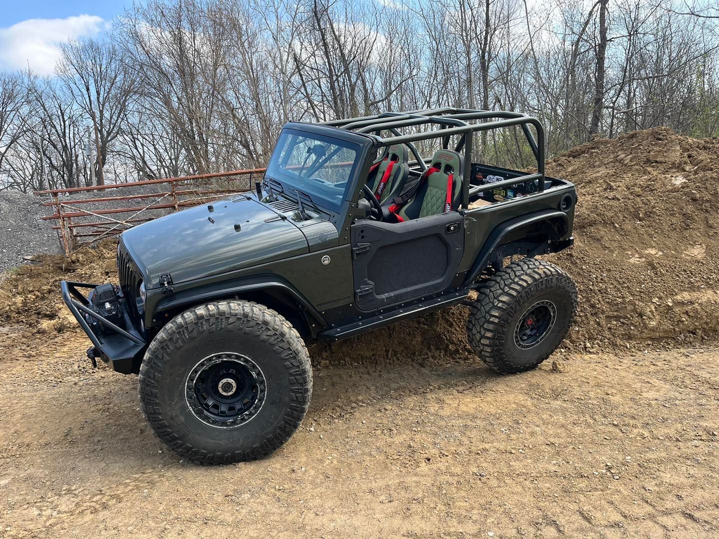 Dark green Jeep Wrangler with large off-road tires and roll cage on a dirt track.