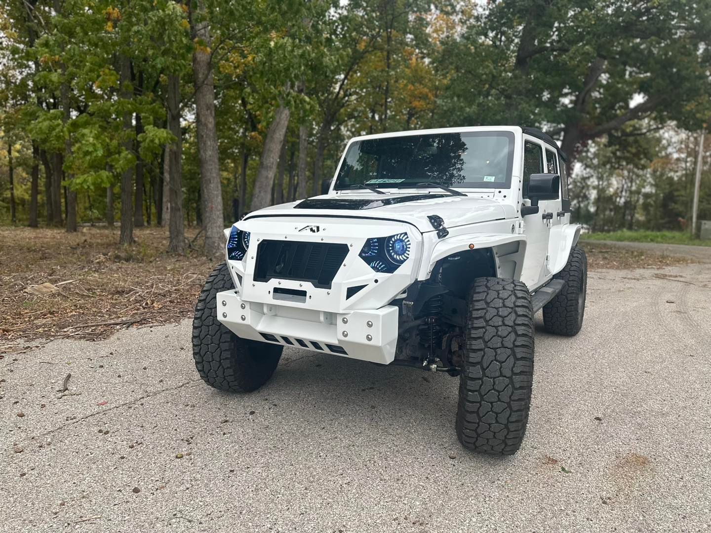 White customized Jeep Wrangler on a gravel road, large off-road tires, modified front grill, forest background.