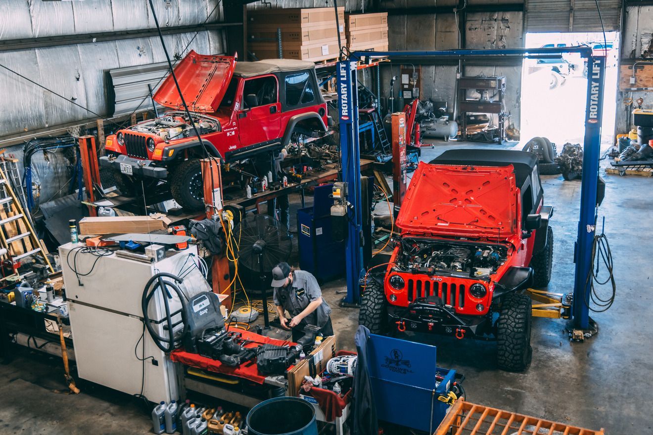 Mechanic working on Jeep in auto shop. Red Jeeps on lifts; tools and parts scattered.