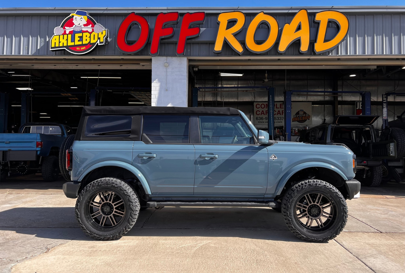 Blue Ford Bronco with black wheels parked in front of an off-road shop.