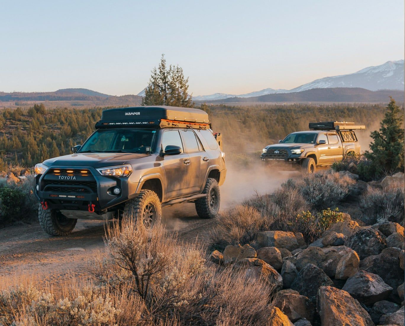 Two off-road SUVs driving on a dirt road in a desert landscape, kicking up dust. Setting sun, mountains in the distance.