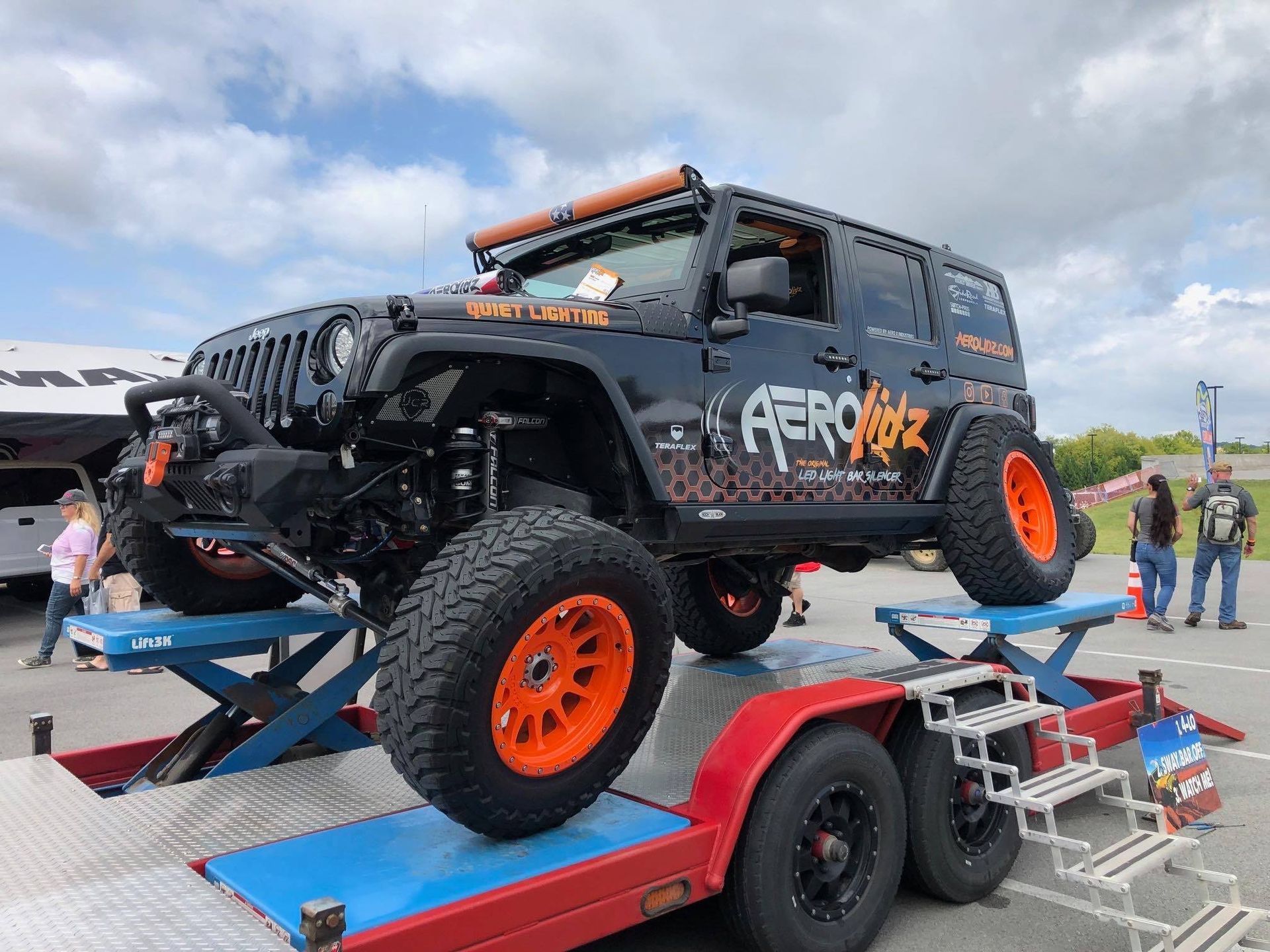 Black Jeep Wrangler with orange accents and large tires on a trailer at an outdoor event.