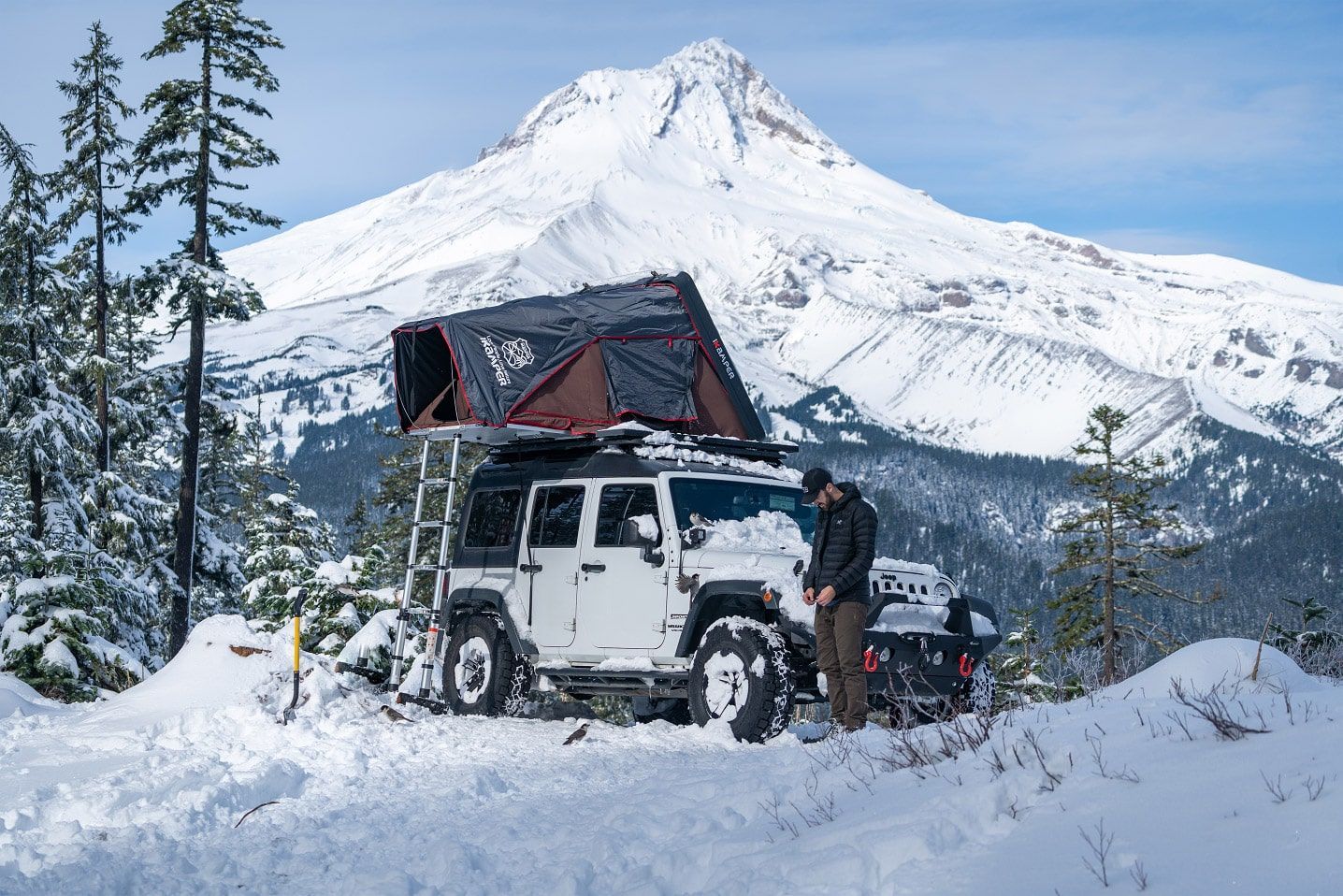 White Jeep with rooftop tent parked in snow, man near it, snow-covered mountain in background.
