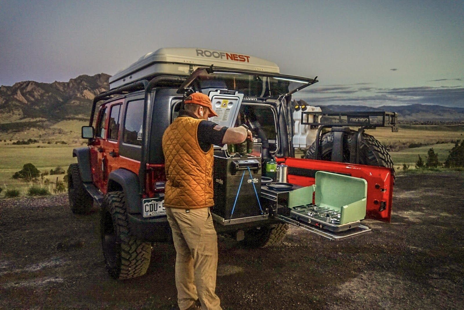 Man camping, setting up a kitchen out of a Jeep's trunk. Orange Jeep, outdoor setting, sunset.