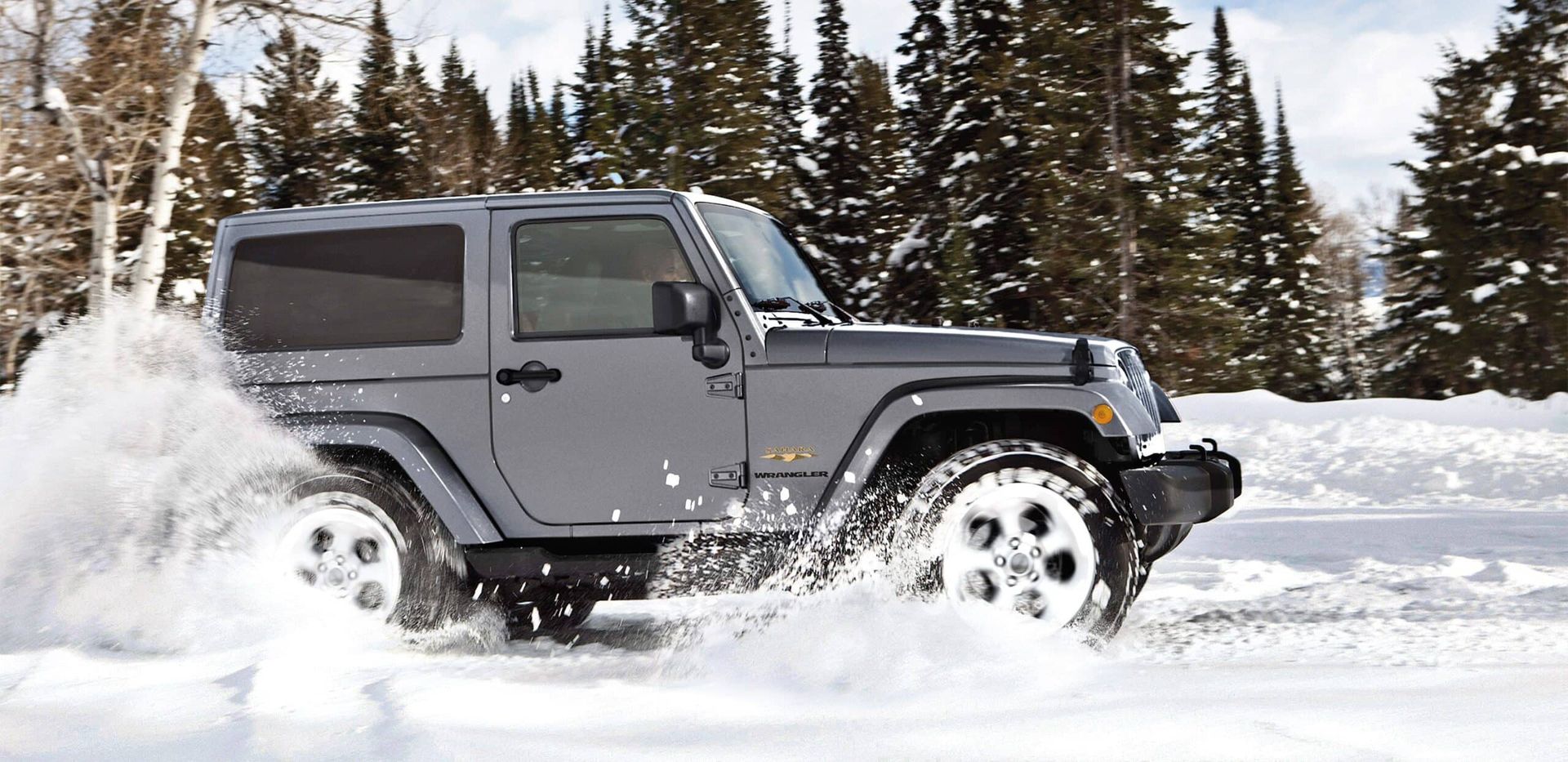 Gray Jeep Wrangler driving through deep snow in a snowy forest.