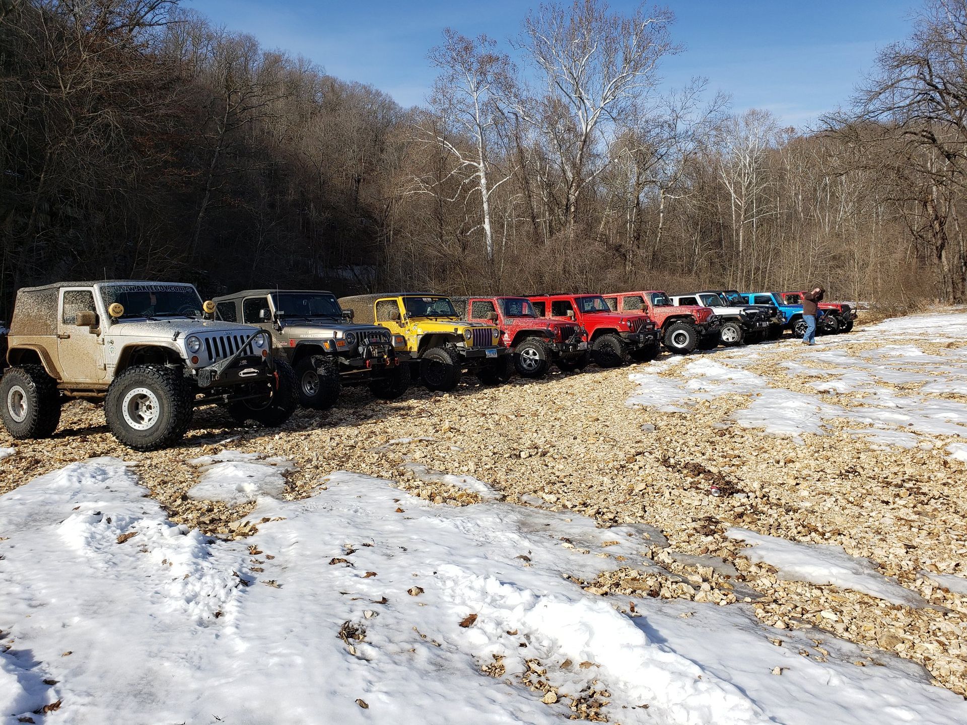 Line of colorful Jeeps parked in snow, along a tree-lined path on a sunny day.