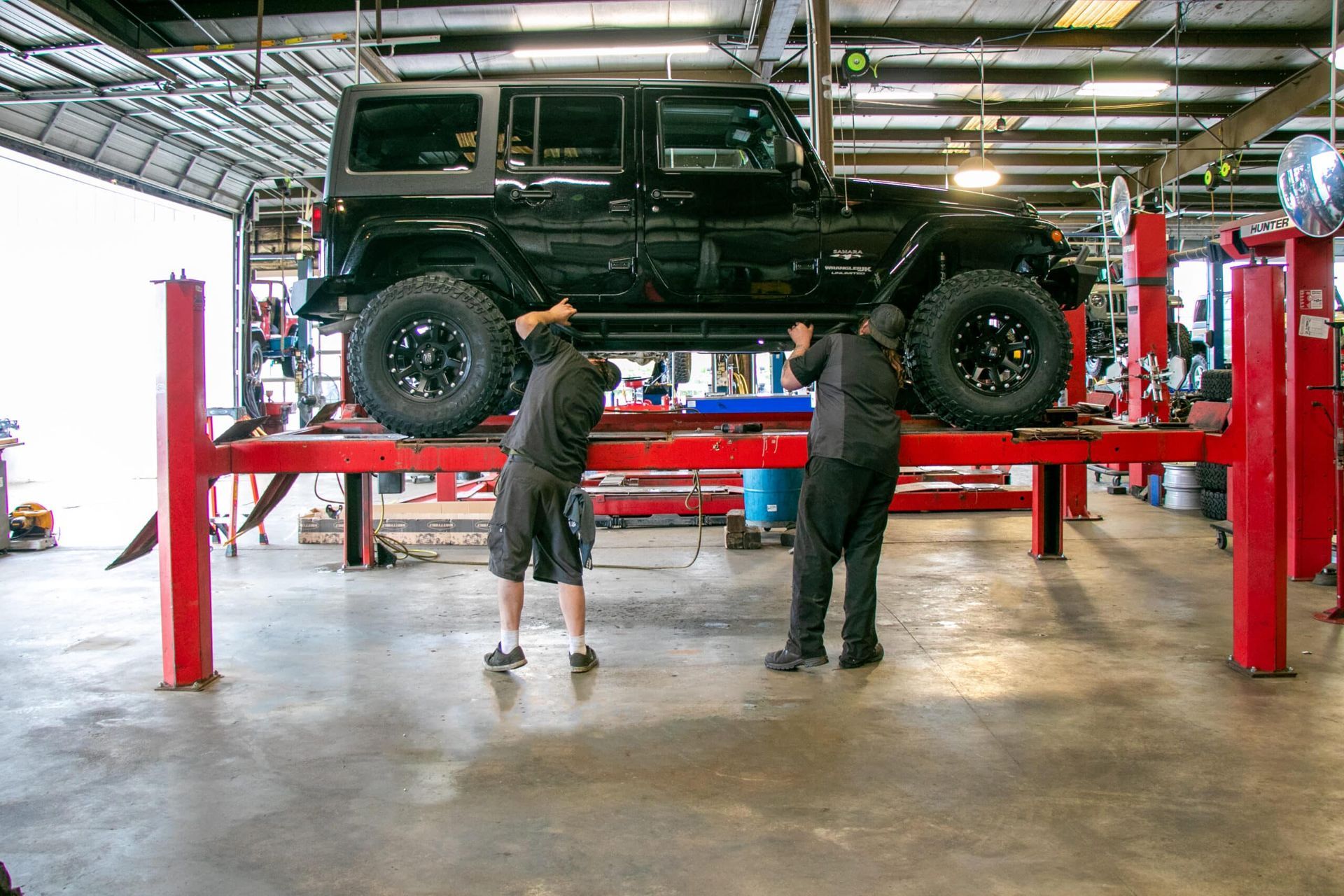 Two mechanics working on a black Jeep lifted on a red hydraulic hoist in a garage.