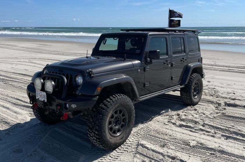 Black Jeep Wrangler parked on a sandy beach, with a roof rack and flag.