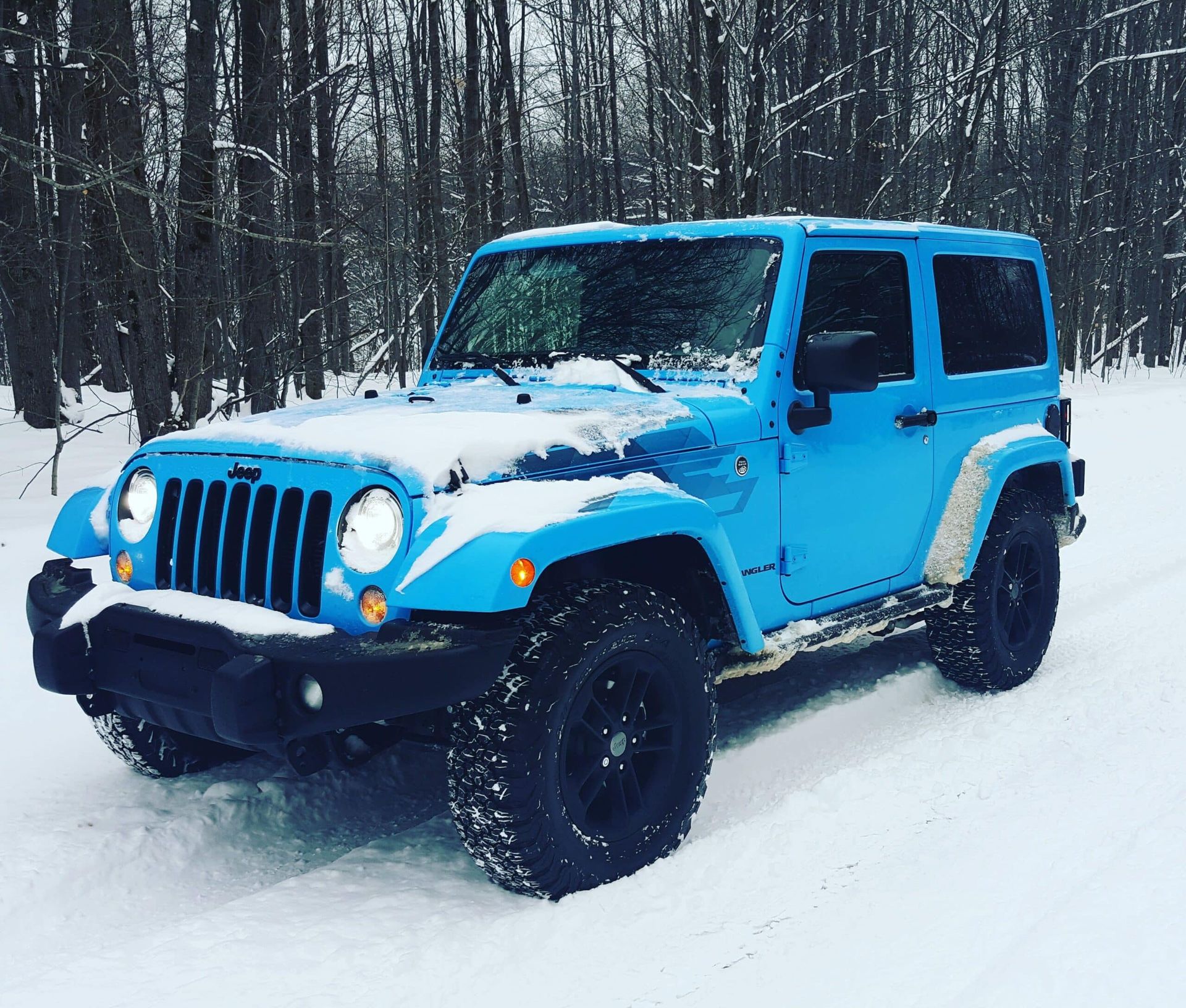 A bright blue Jeep Wrangler driving on a snowy forest road.