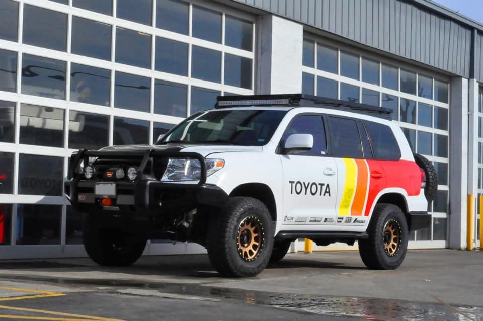 White Toyota Land Cruiser with black accents, roof rack, and colorful side graphic, parked in front of a garage.