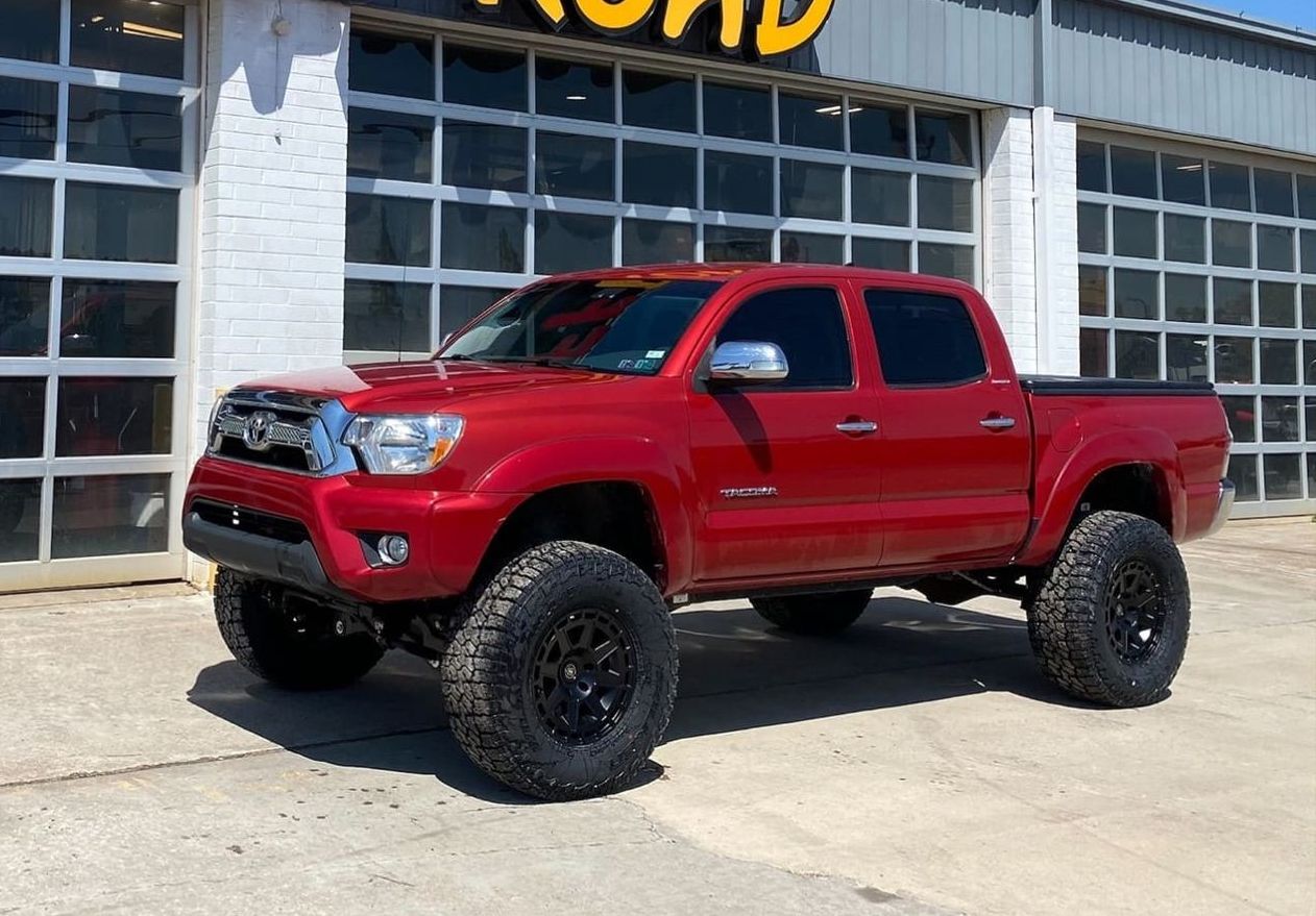 Red lifted Toyota Tacoma truck with black wheels parked in front of a shop.