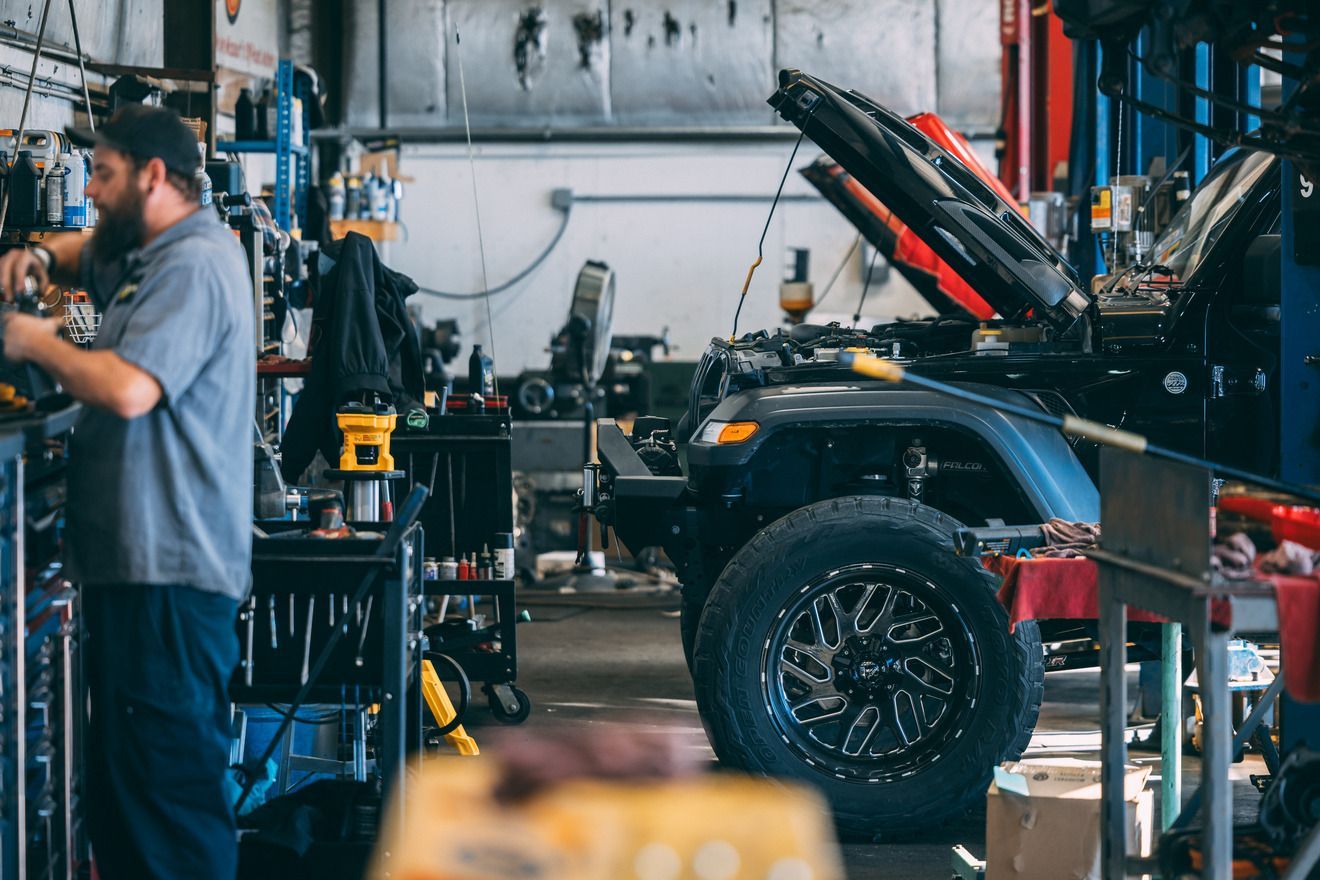 Mechanic working on a dark-colored Jeep in a cluttered auto repair shop.
