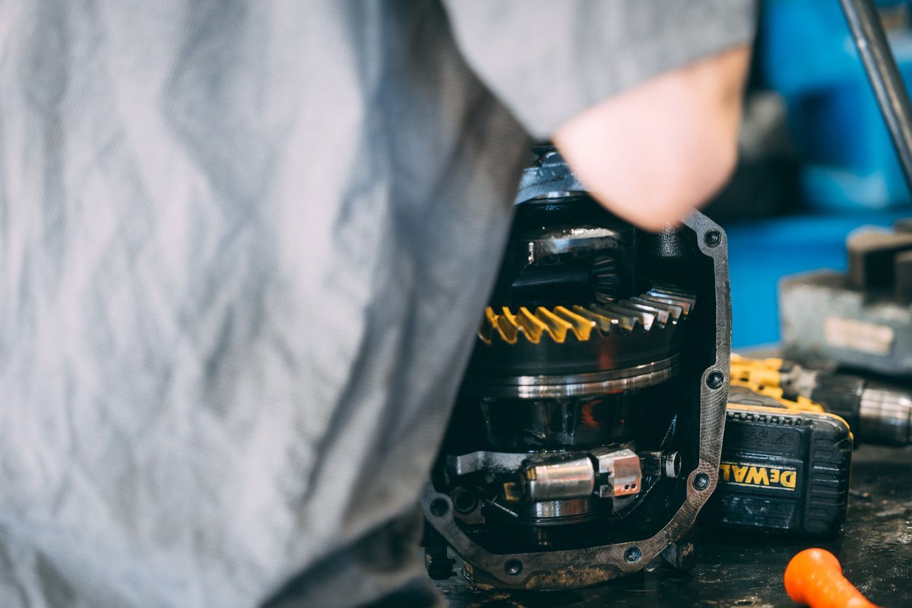 Mechanic working on a differential gear assembly. A yellow gear visible with tools and a drill in the shop.
