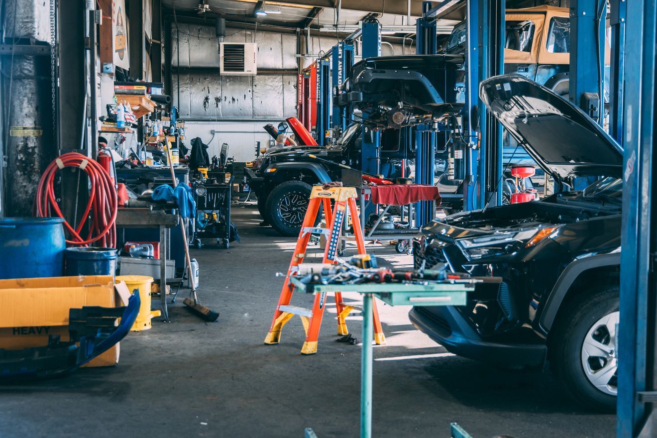 A cluttered auto repair shop with cars on lifts, tools, and a ladder.