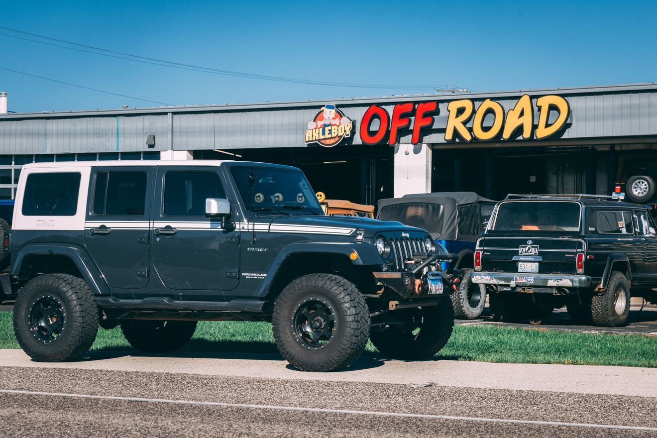Jeep Wrangler in front of an off-road shop; gray SUV with large tires, black rims, and a white top.