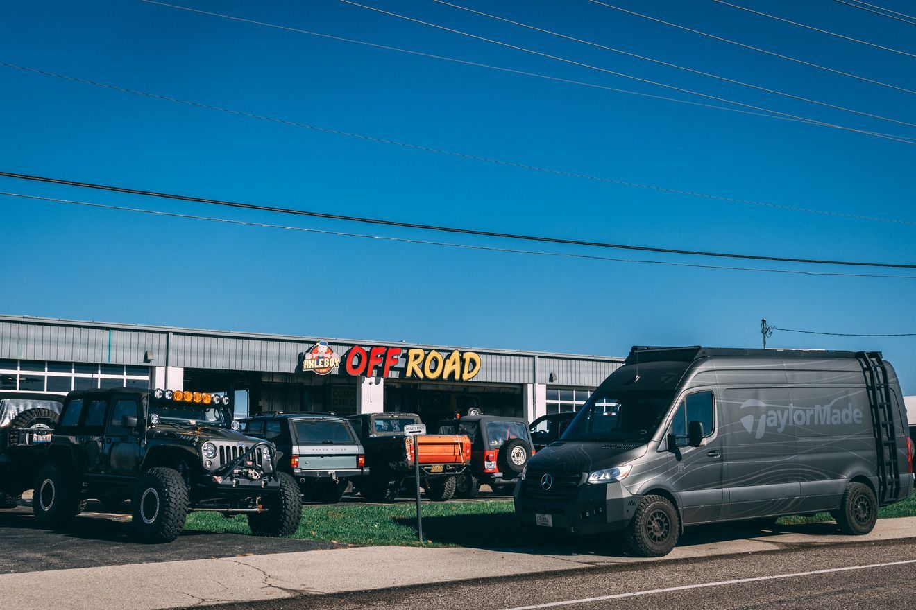 Off-road vehicle shop with a van and several customized trucks parked in front on a sunny day.