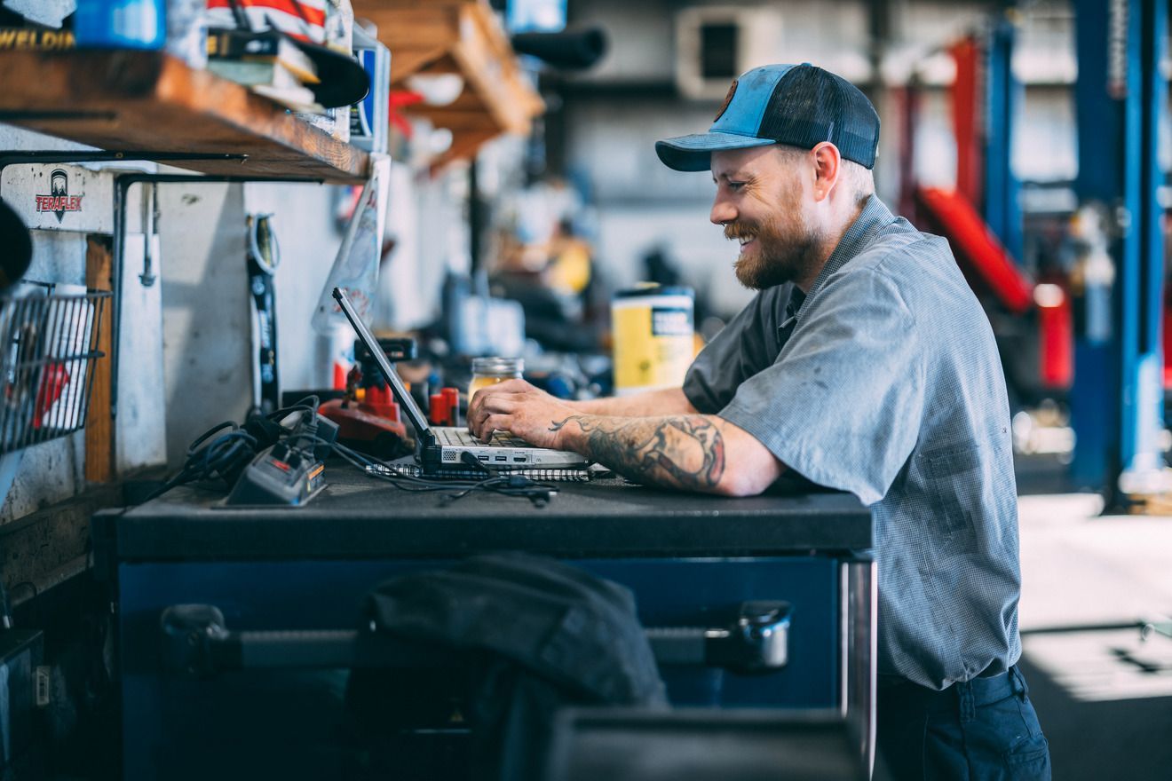 Mechanic smiling while using a laptop at a workbench in a cluttered garage.