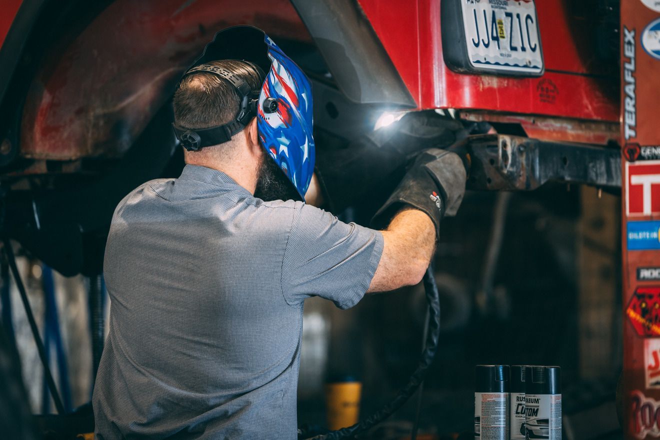 Person welding a red car with a blue and white star-patterned mask, in a garage setting.