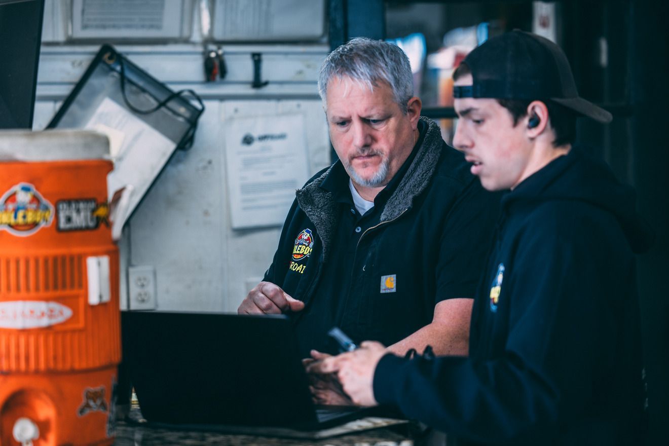 Two men looking at a laptop, one older with gray hair and one younger with a cap, in an office setting.