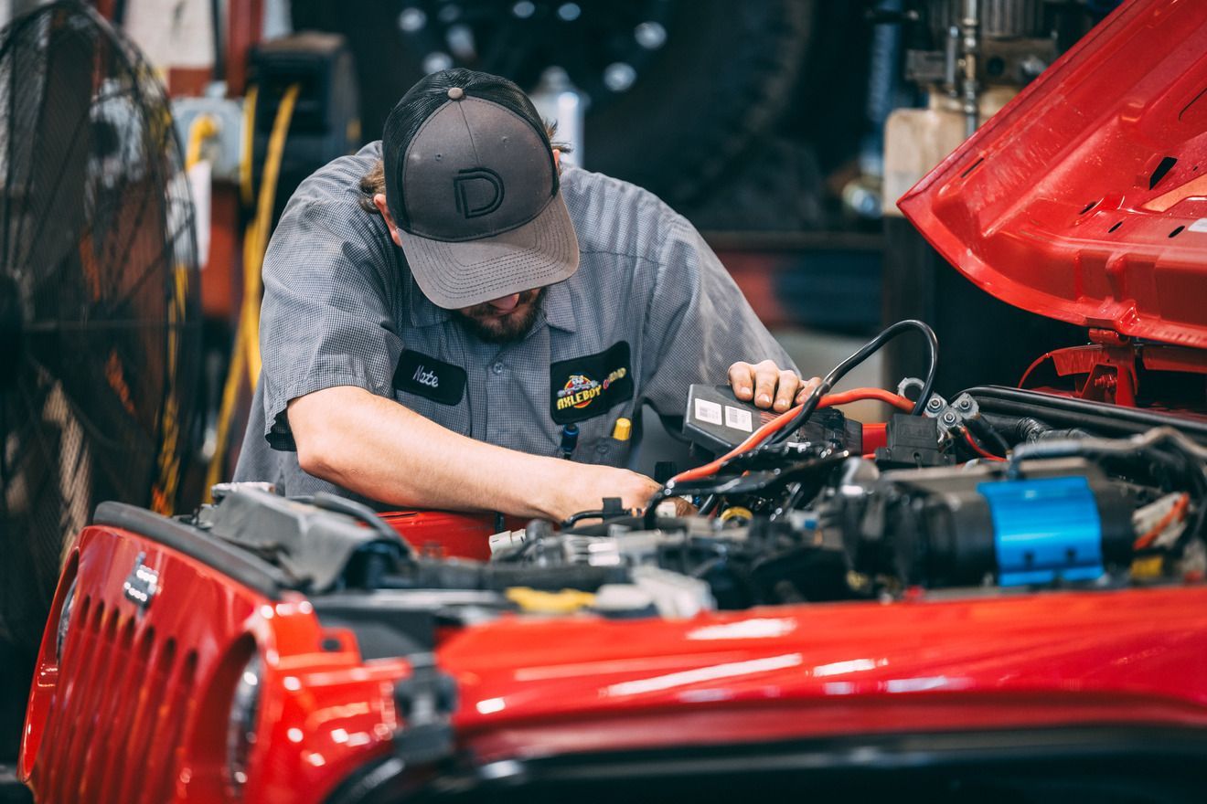 Mechanic working on the engine of a red Jeep. Wearing a hat and uniform, inside a garage.
