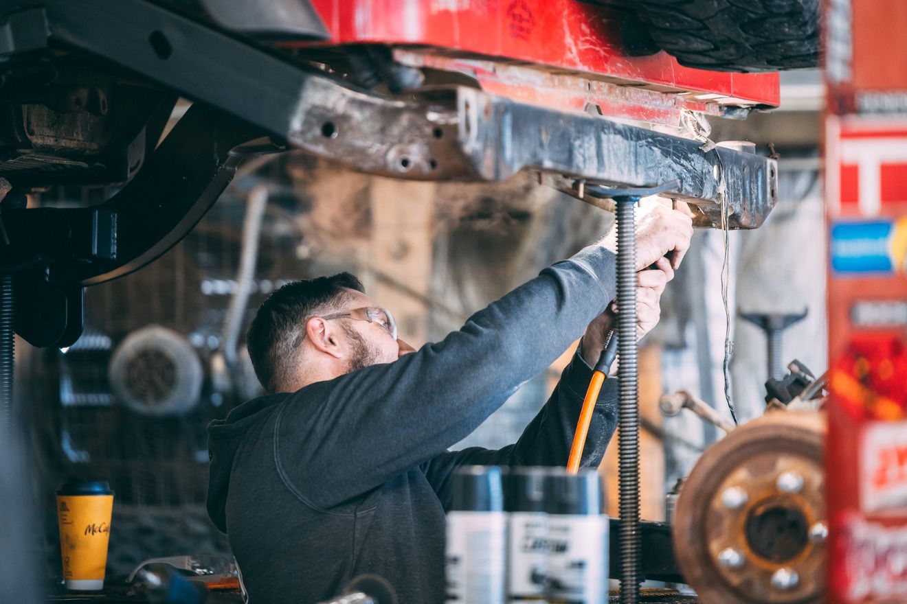 Mechanic working on a car's undercarriage in a garage. He wears glasses, and uses tools.