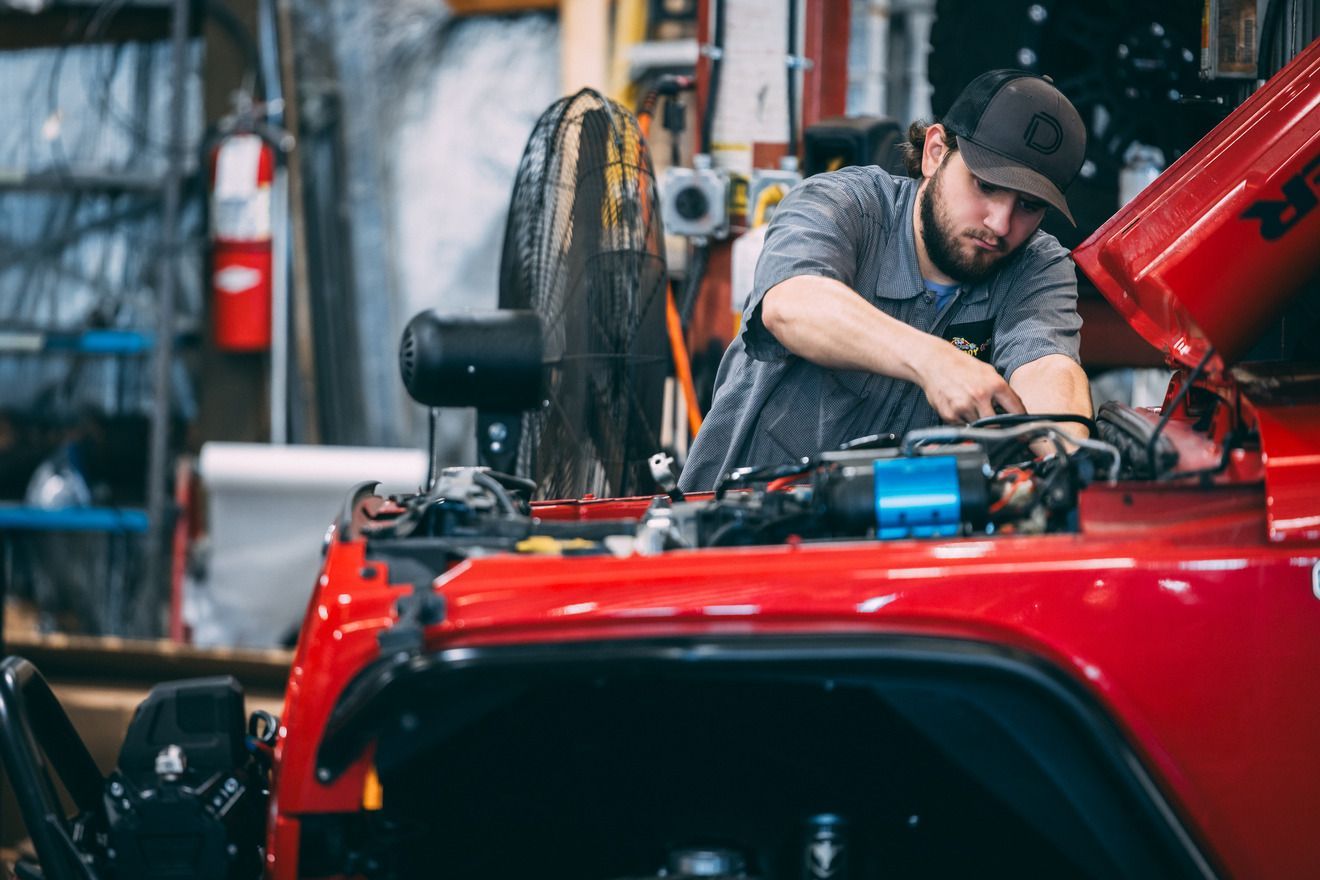 Mechanic working on a red Jeep engine.  He wears a cap and gray shirt in a garage.