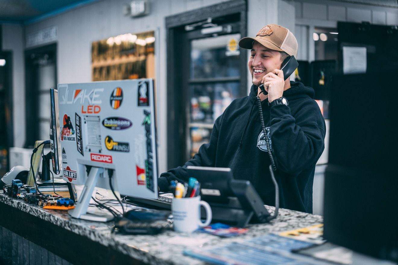 A smiling person on the phone at a front desk. They are wearing a hat and hoodie, with stickers on the monitor.
